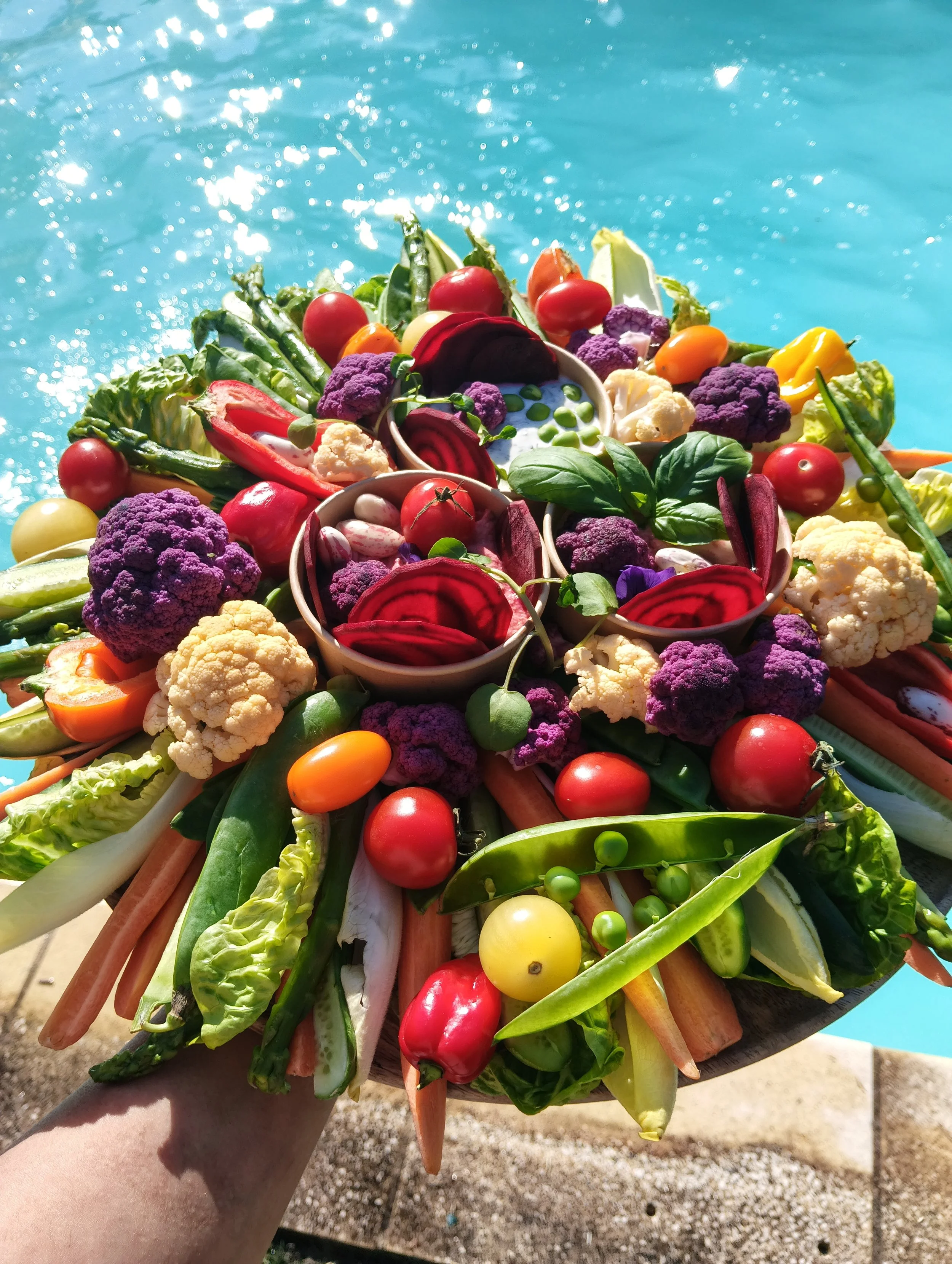 Présentation colorée de légumes variés comme tomates, choux, radis, poivrons, asperges, et autres, disposés sur une tablette au bord d'une piscine.