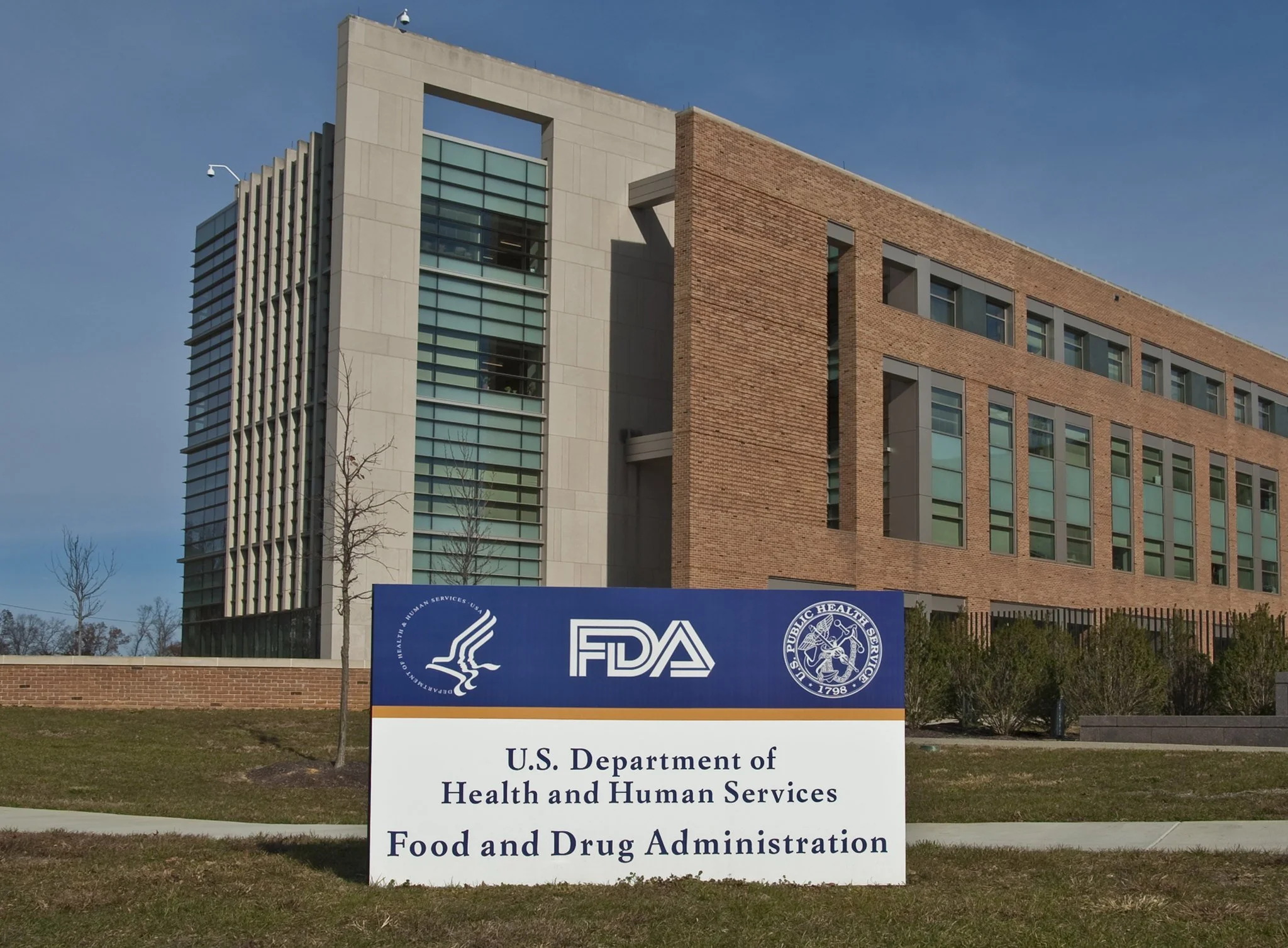 A building with a sign that reads U.S. Department of Health and Human Services and Food and Drug Administration in the foreground, with a modern, multi-story brick and glass structure behind it under a clear blue sky.