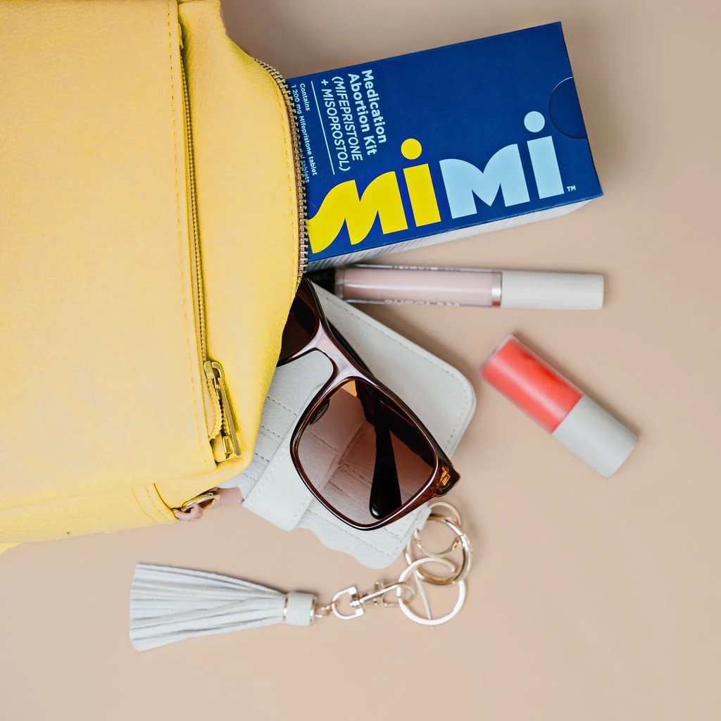 Travel essentials including a yellow zippered pouch, a box of an over-the-counter medication abortion prototype, a tube of lip balm, sunglasses in a white case, and a keychain with a white tassel.