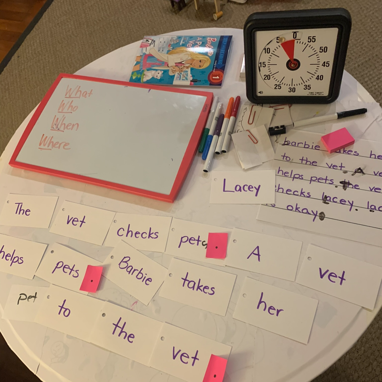 Educational workspace with a whiteboard, learner's writing, colored markers, paper clips, and sentences related to a story about Barbie. A large timer or clock and a children's magazine are also on the table, which is covered with educational materials and word cards.