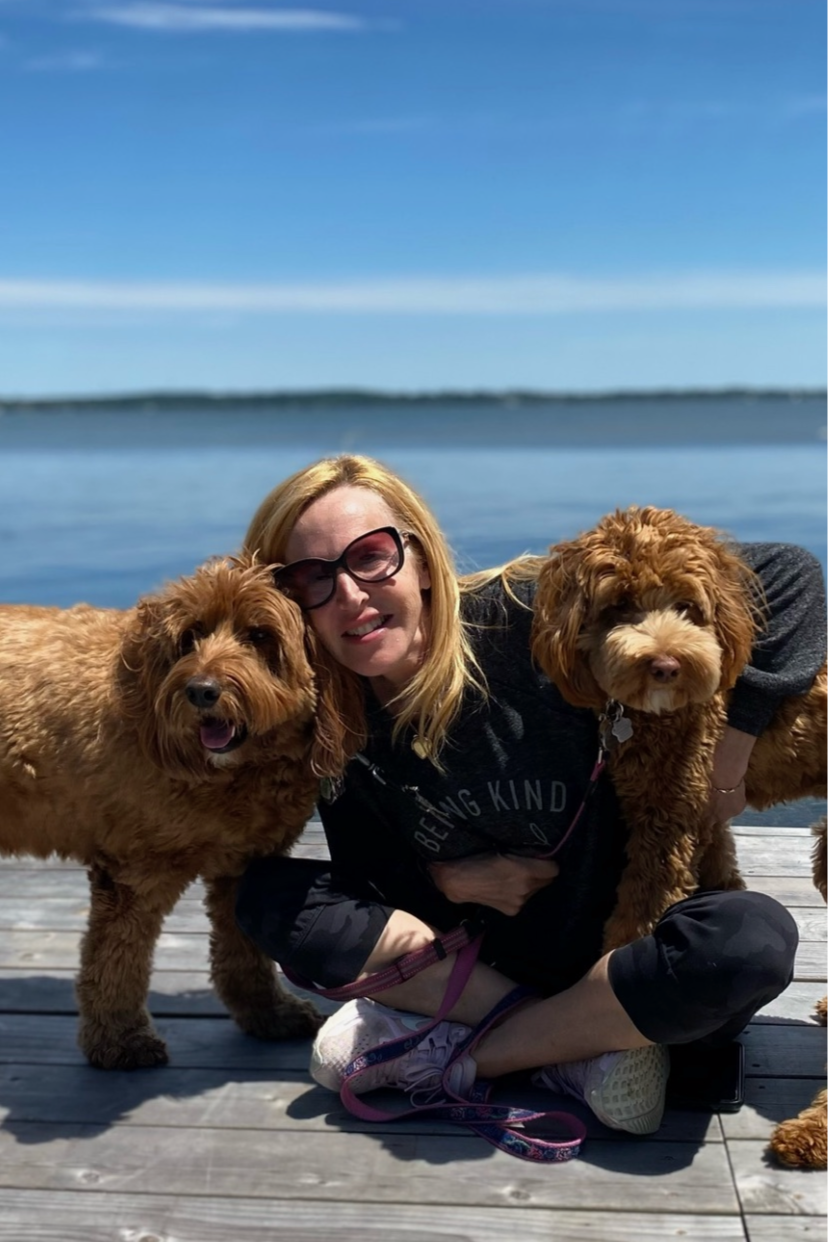 Woman with sunglasses sitting on a wooden dock by a lake, surrounded by two brown fluffy dogs, with a blue sky and water in the background.