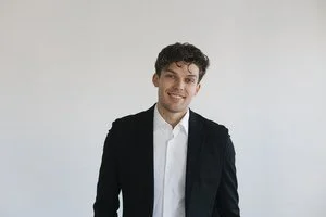 A young man in a black blazer and white shirt smiling against a plain light background.
