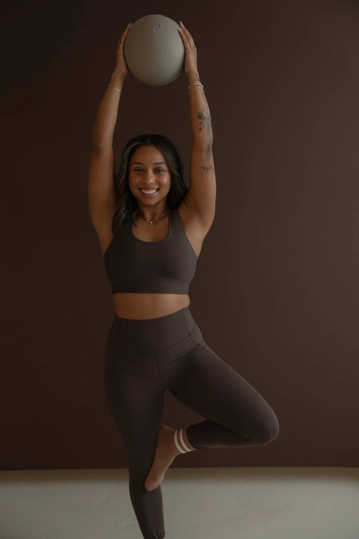 A woman performing a yoga pose with one leg raised, balancing on her other leg, holding a large exercise ball overhead, wearing black athletic wear and smiling.
