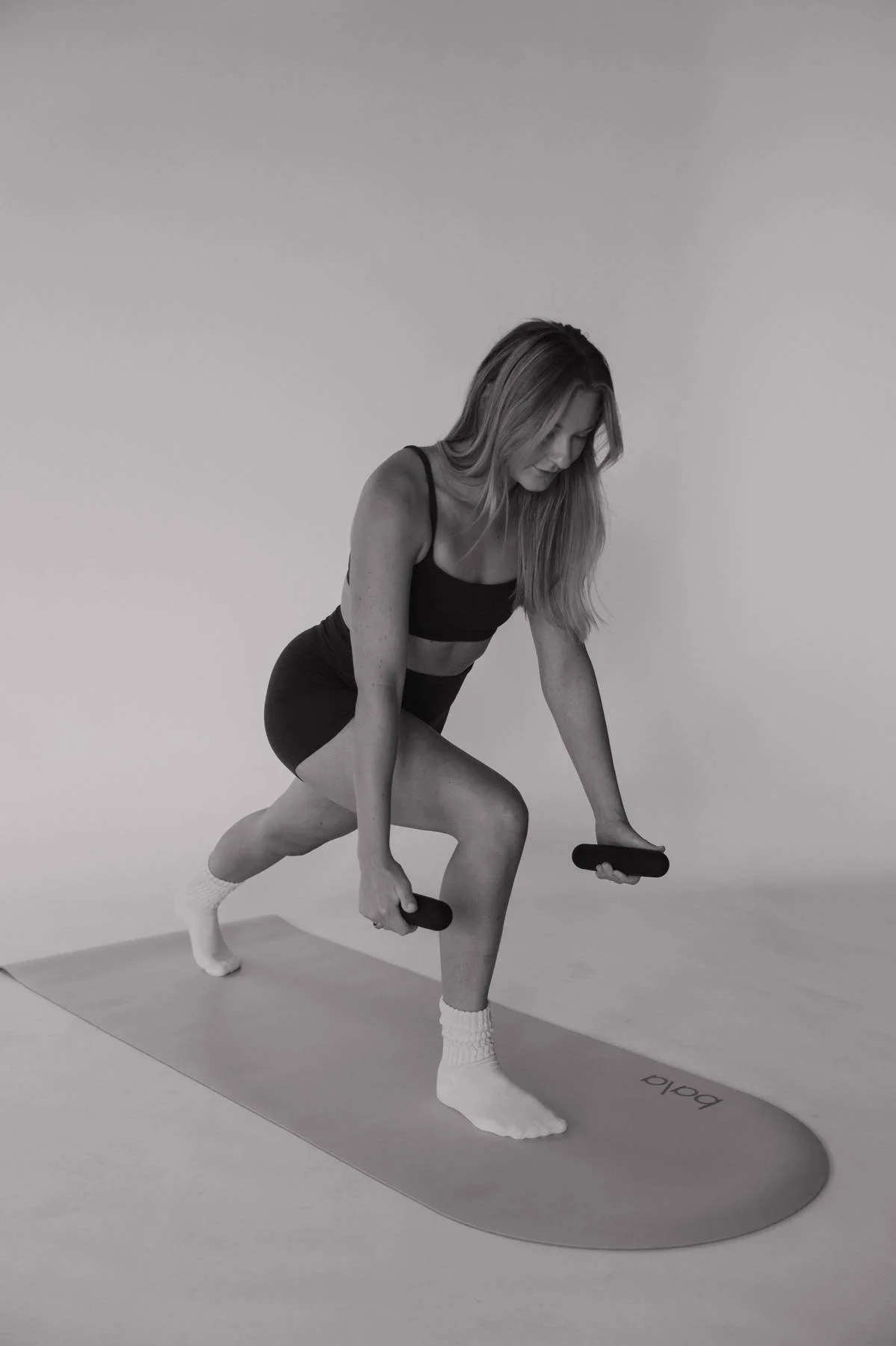 Black and white photo of a woman in athletic clothing doing a lunge exercise on a yoga mat, holding dumbbells in both hands, against a plain background.