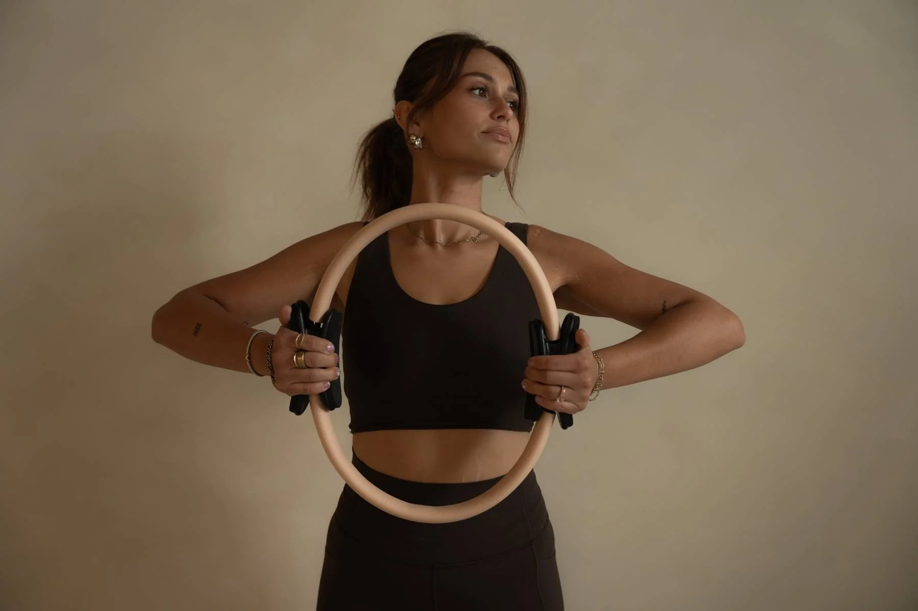 A young woman holding a yoga or fitness ring in front of her chest, wearing a black sports bra and black leggings, standing against a plain beige wall.