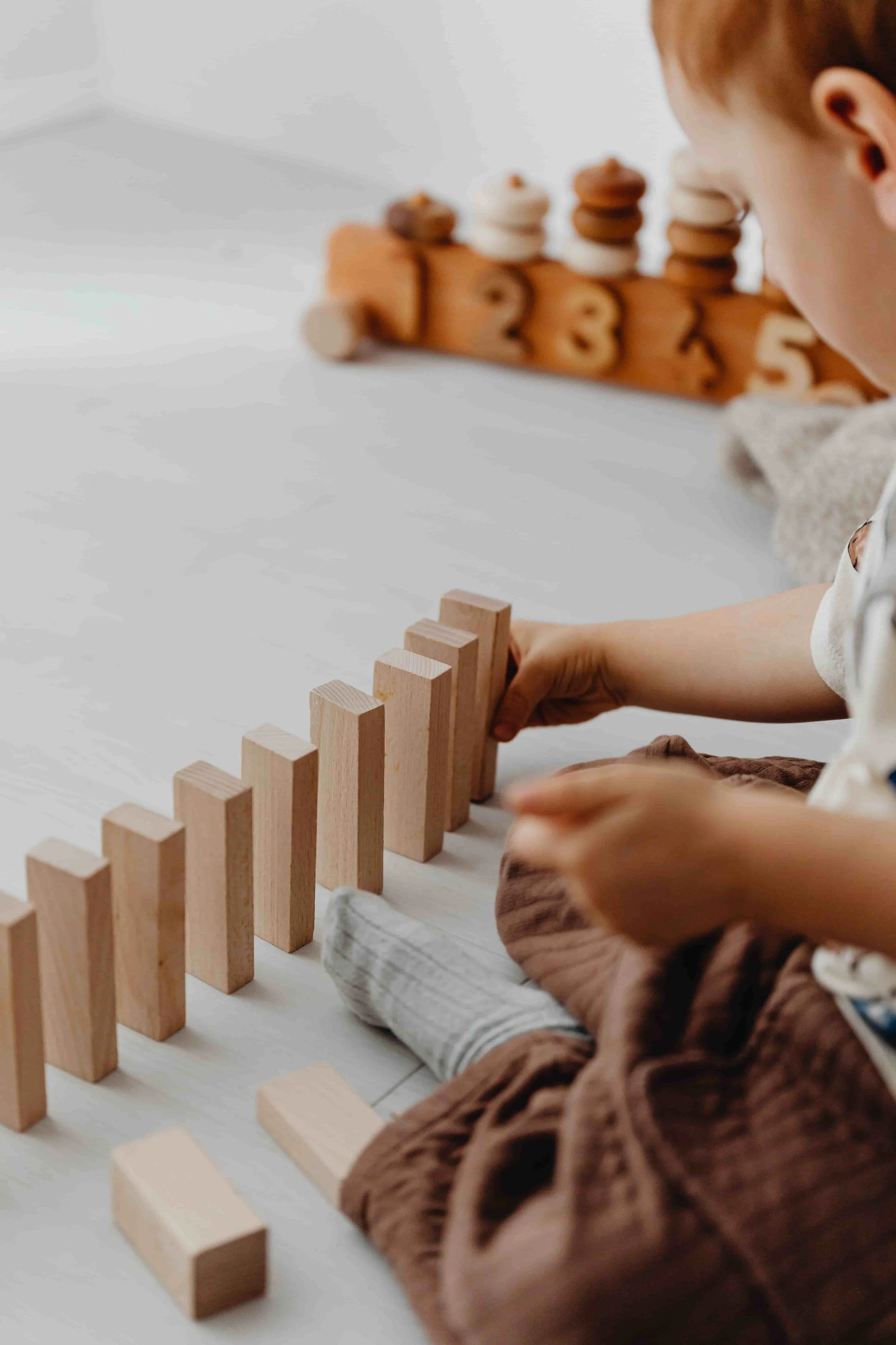 A child playing with wooden toys, stacking blocks on a white surface with a wooden rack holding numbered miniature figure pieces in the background.