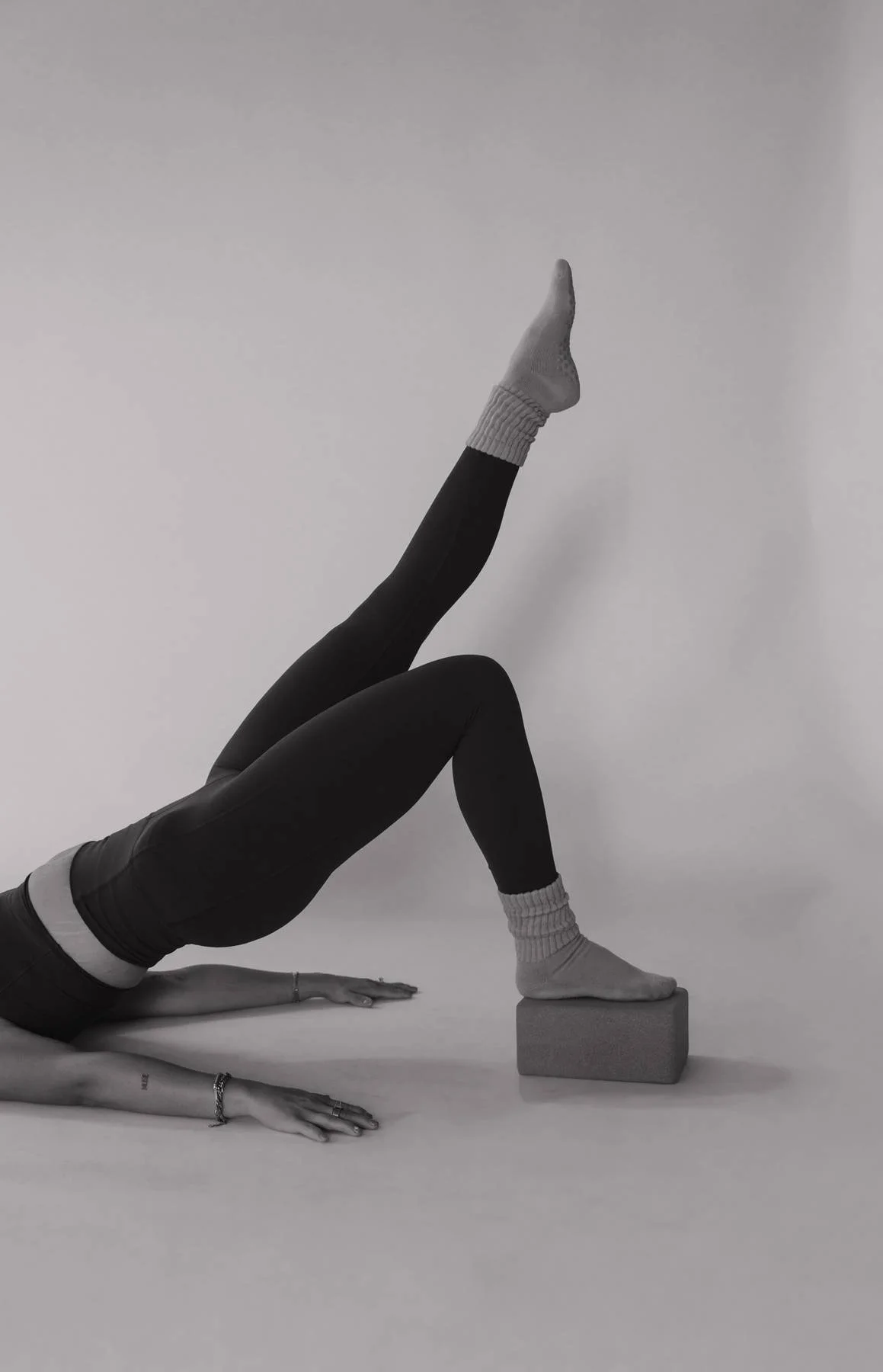 A person lying on the floor with one leg raised and supported by a foam block, wearing black leggings, socks, and a long-sleeve shirt, against a plain background.