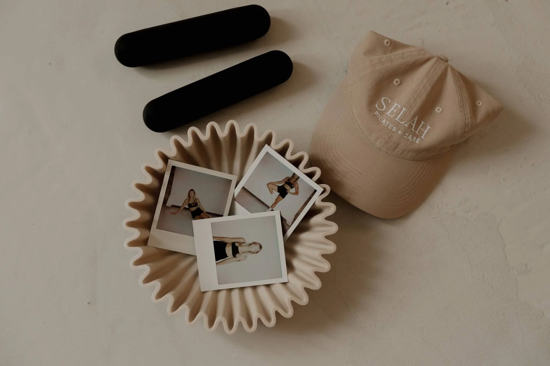 A collection of items on a white surface including a beige hat with 'SELAH Pilates & Care' embroidered on it, a beige ruffled dish holding three Polaroid photos of a woman in black workout clothes, and two black cylindrical objects.