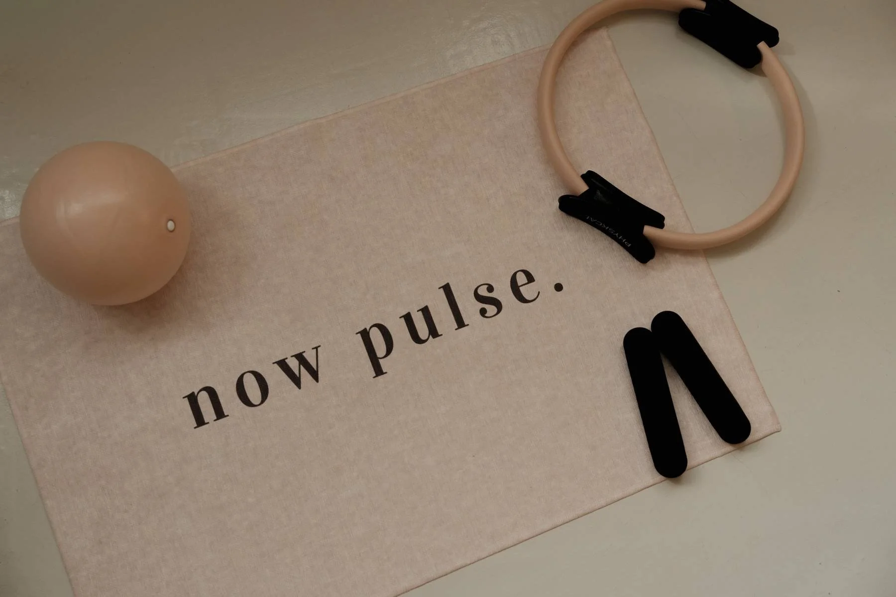 A beige sphere, a pink resistance band with black handles, black letters 'M', and a paper with the text 'now pulse.' on it, arranged on a flat surface.