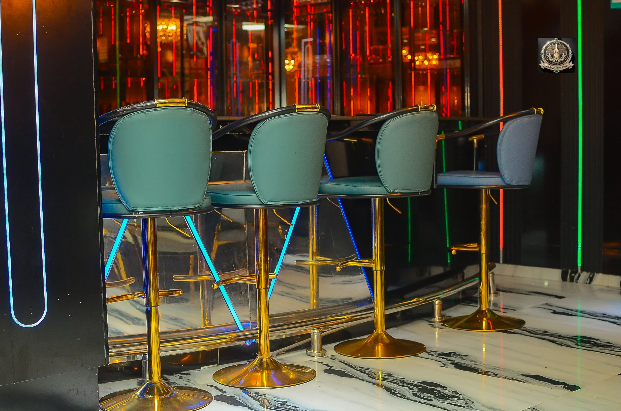 A row of teal bar stools with gold bases clustered in front of a neon-lit wall in a modern bar or nightclub.