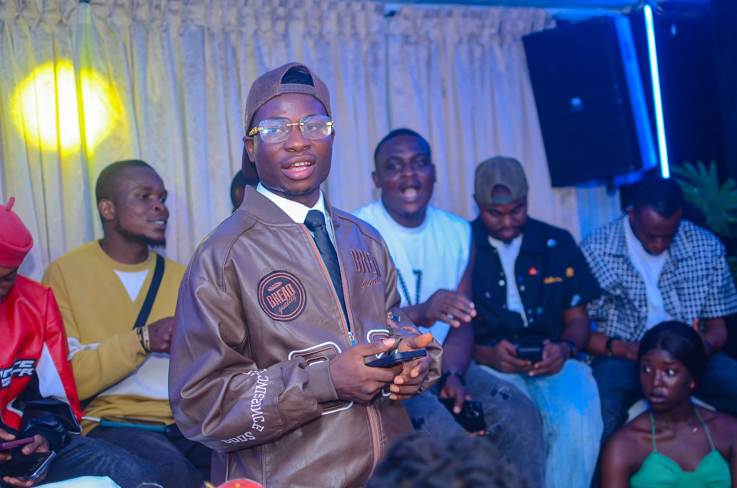 A group of young men sitting indoors, with one man standing in front of them holding a phone. The man standing is wearing a brown jacket, glasses, and a cap worn backwards. The background features a curtain and blue lighting.