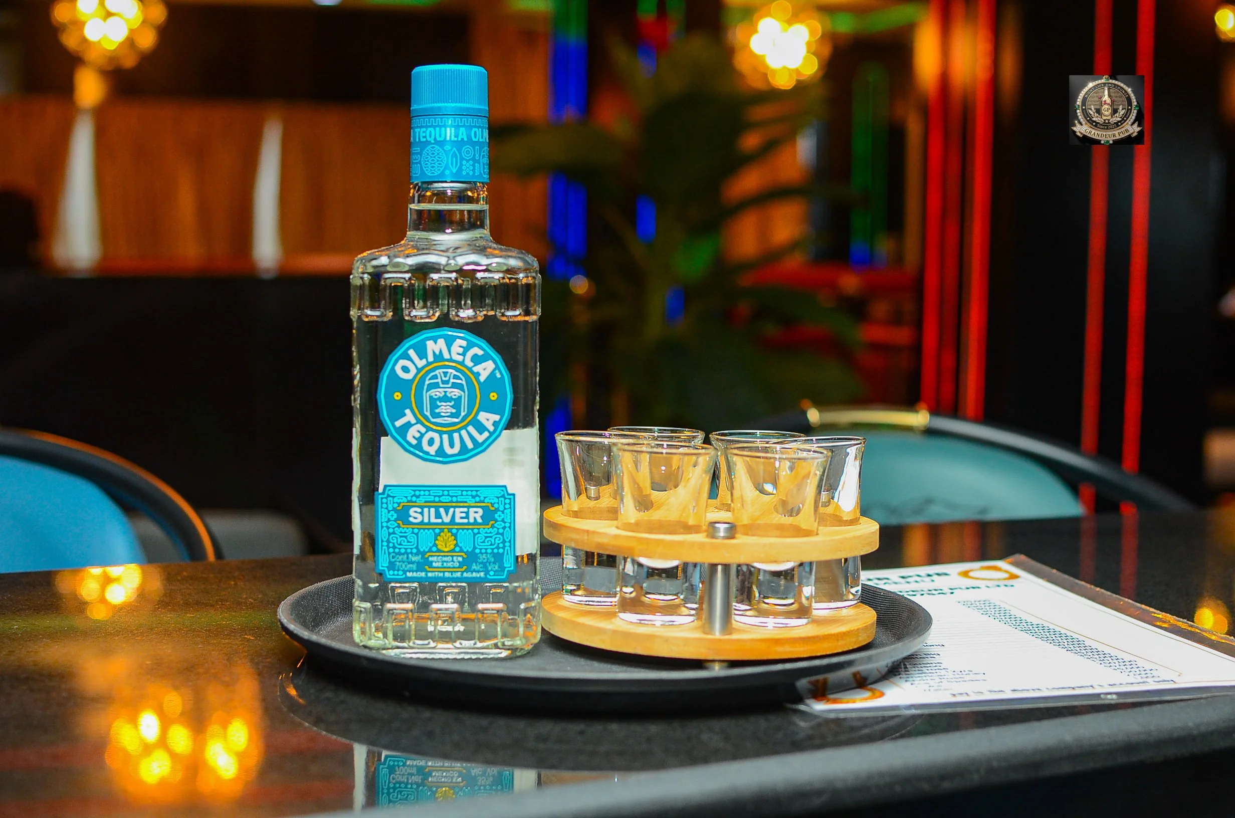 A bottle of Olmeca Silver tequila on a black tray with shot glasses, set on a table in a dimly lit bar or restaurant with colorful lights in the background.