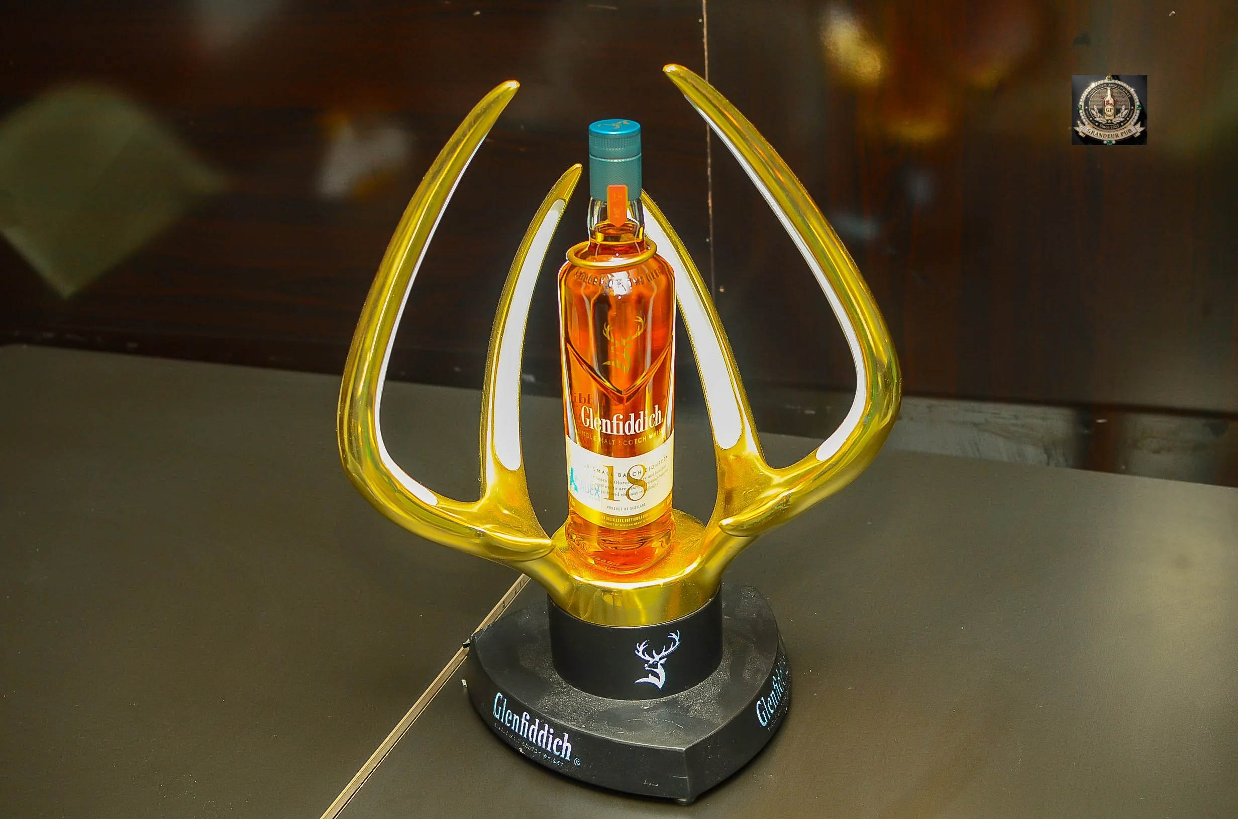 A glass bottle of Glenfiddich single malt Scotch whisky displayed in a decorative yellow and white holder with a black base branded with the Glenfiddich logo, placed on a black surface with a dark background.