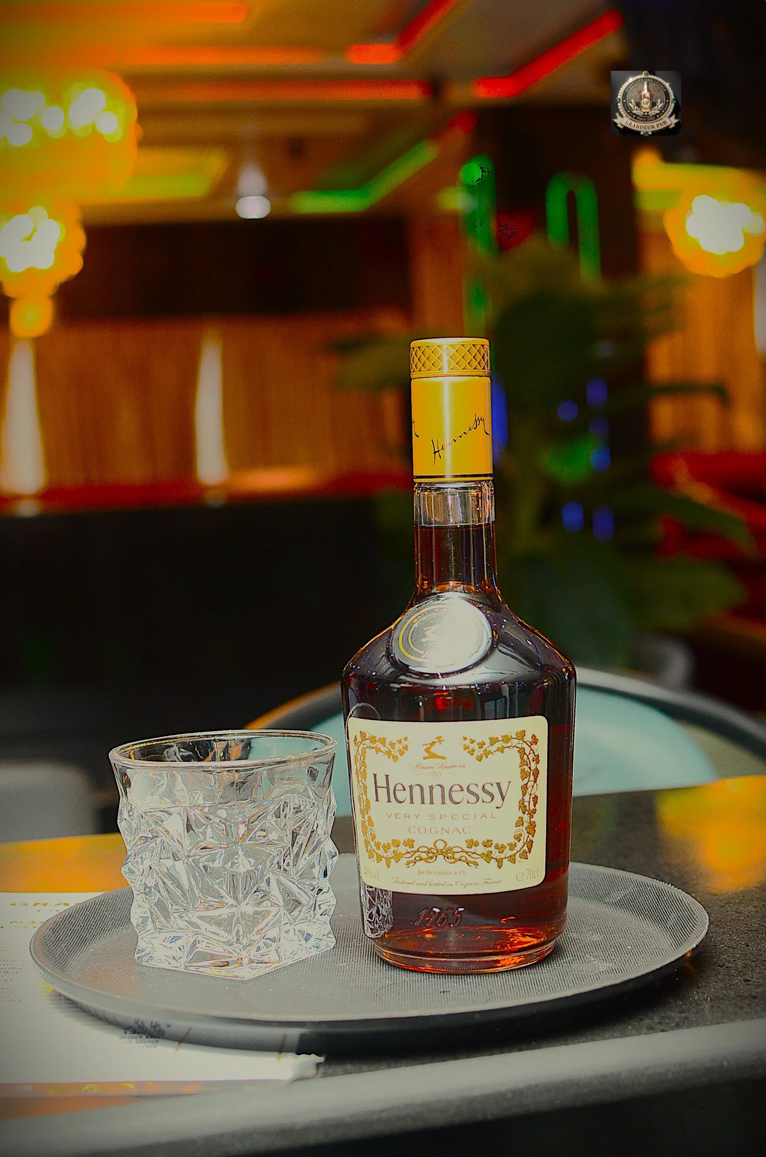 A bottle of Hennessy Very Special Cognac on a silver tray with a crystal glass nearby, set on a dining table with a place setting. The background has colorful blurred lights.
