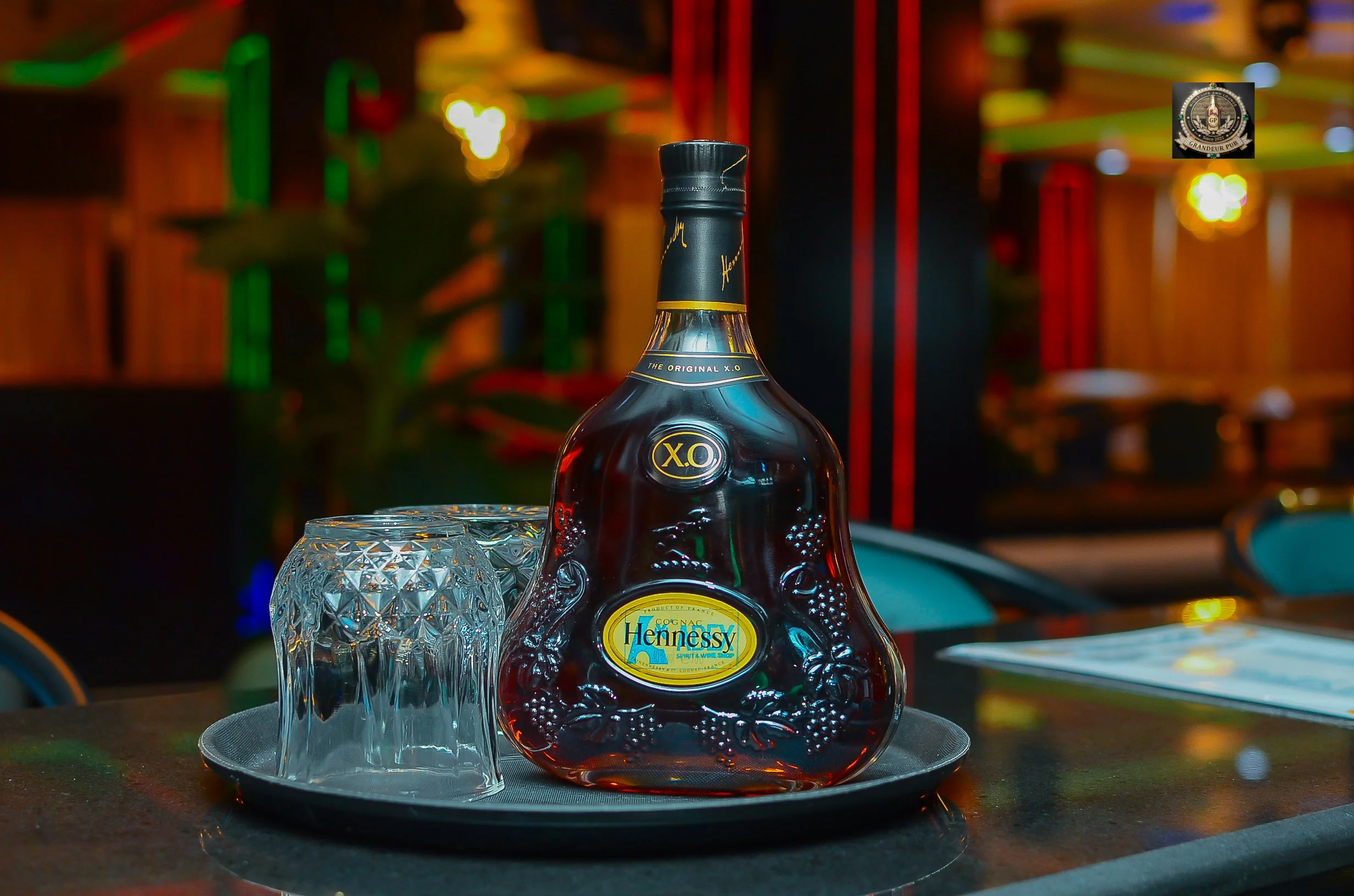 A bottle of Hennessy Cognac next to a glass on a black tray, with a blurred colorful background.