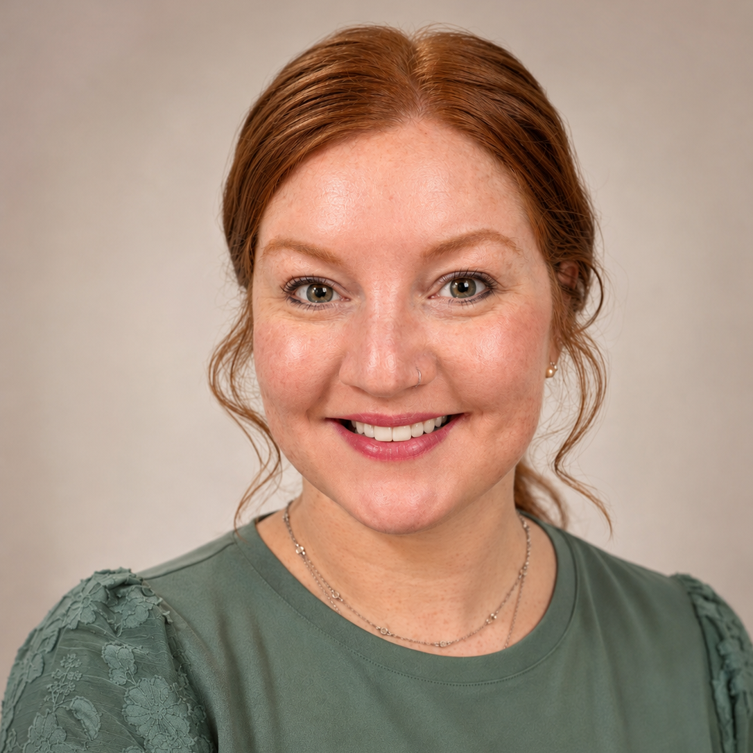 A woman with red hair, light skin, and gray eyes, smiling, wearing a green top and layered necklaces, against a plain background.