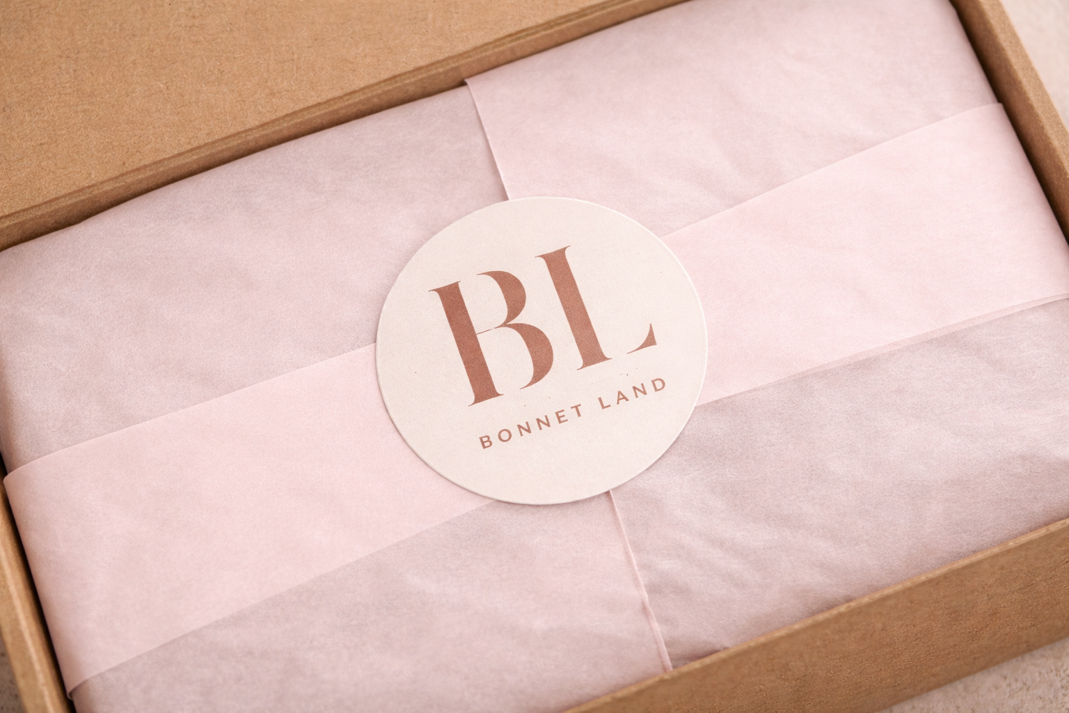 A cardboard box contains neatly wrapped pink tissue paper with a round white sticker featuring the initials 'BL' and the text 'Bonnet Land'.