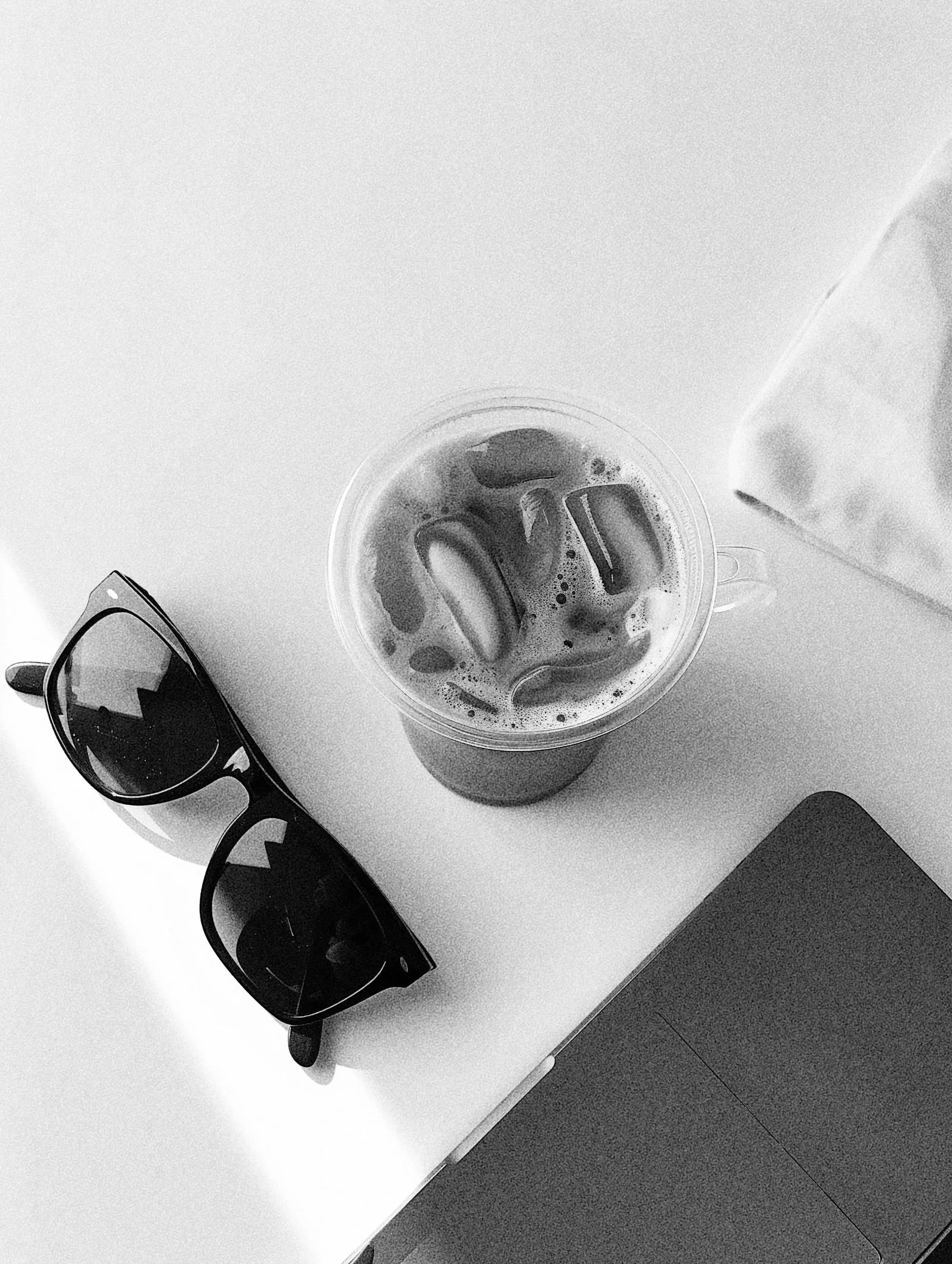 A drink with ice cubes in a clear plastic cup, placed on a white desk with a pair of dark sunglasses and a wallet nearby, alongside a disposable cup, with the image taken from above in black and white.