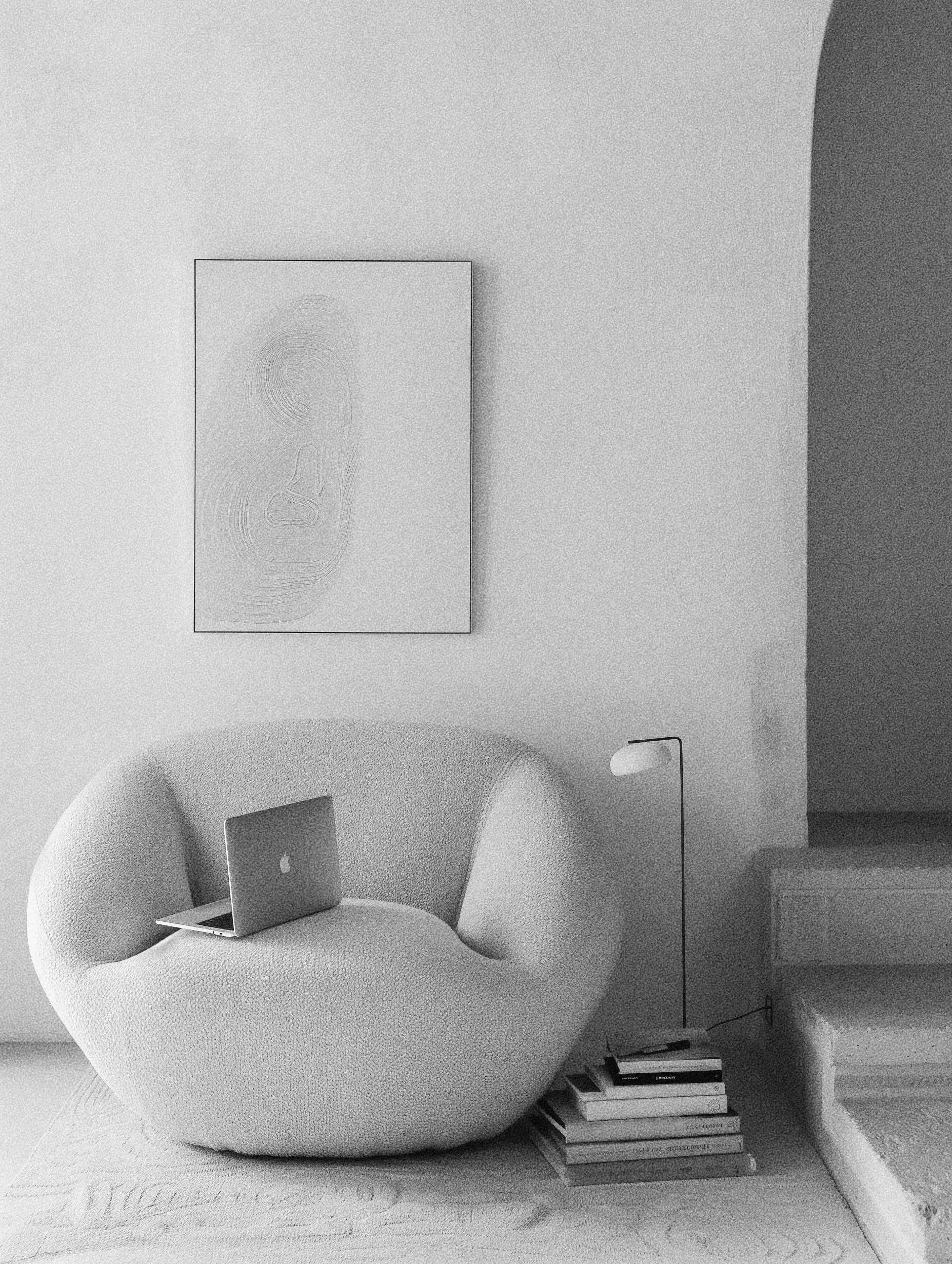 A curved, modern armchair with a textured fabric surface, with an open MacBook on the seat. Next to the chair, there is a small table with a stack of books underneath. The background includes a wall with framed artwork and a shelf with books.