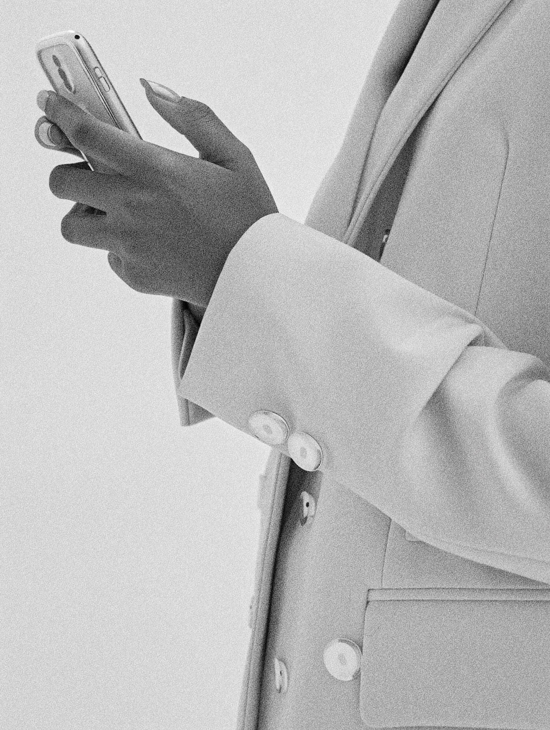 A person wearing a light-colored blazer holding a smartphone in one hand and a stylus or pen in the other, with part of their upper body and arm visible, standing beside a wall with a door frame.
