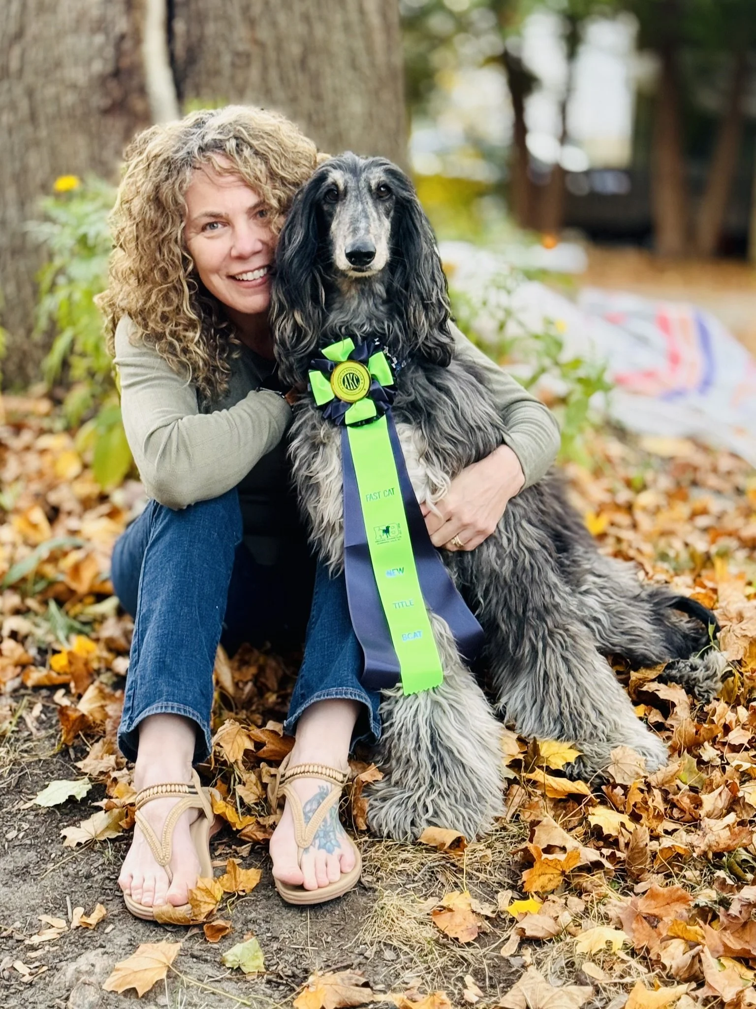 A woman with curly blonde hair smiling and hugging a long-haired dog with a blue and green award ribbon on its neck, sitting on fallen autumn leaves in a park.