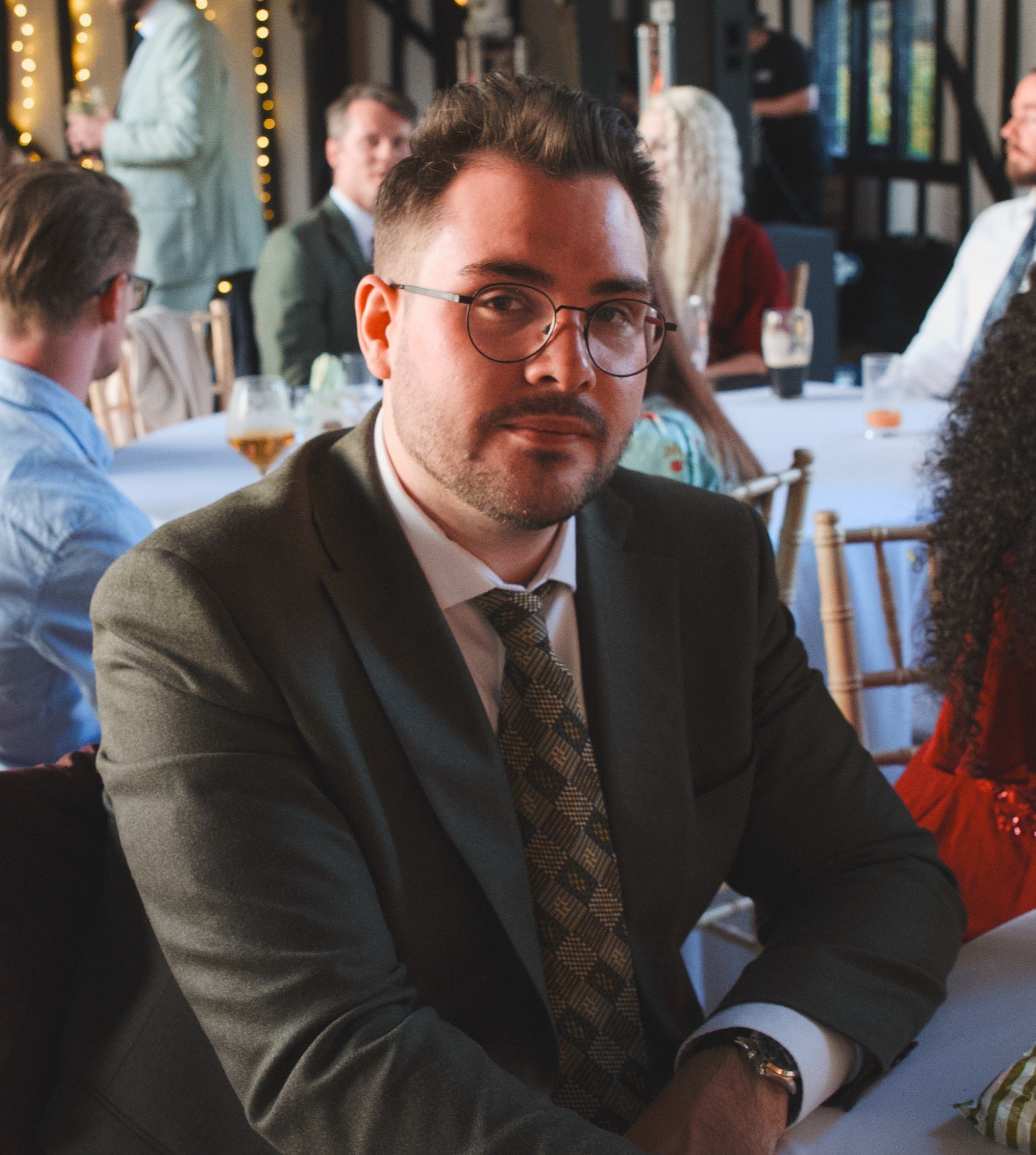 A man wearing glasses and a suit, sitting at a table during a social gathering or celebration, with other people in the background.