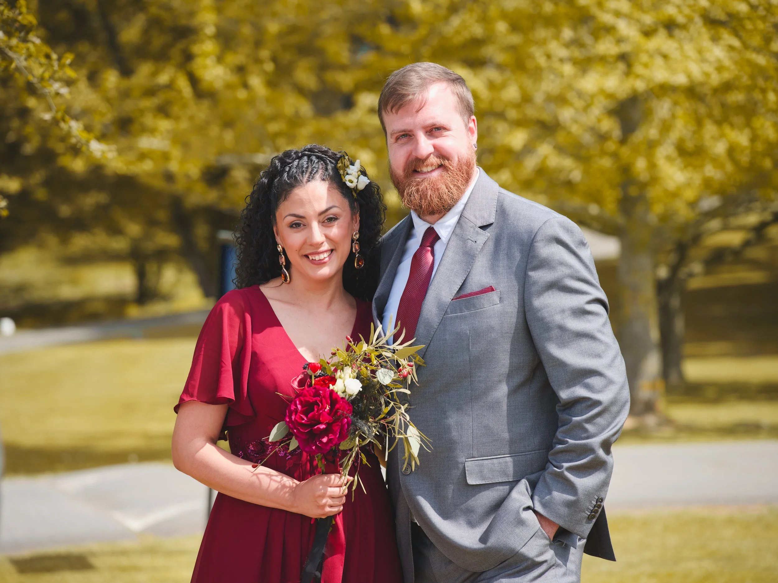 A smiling woman and man in formal attire standing outdoors, holding a bouquet of red and white flowers, with trees and yellowing leaves in the background.