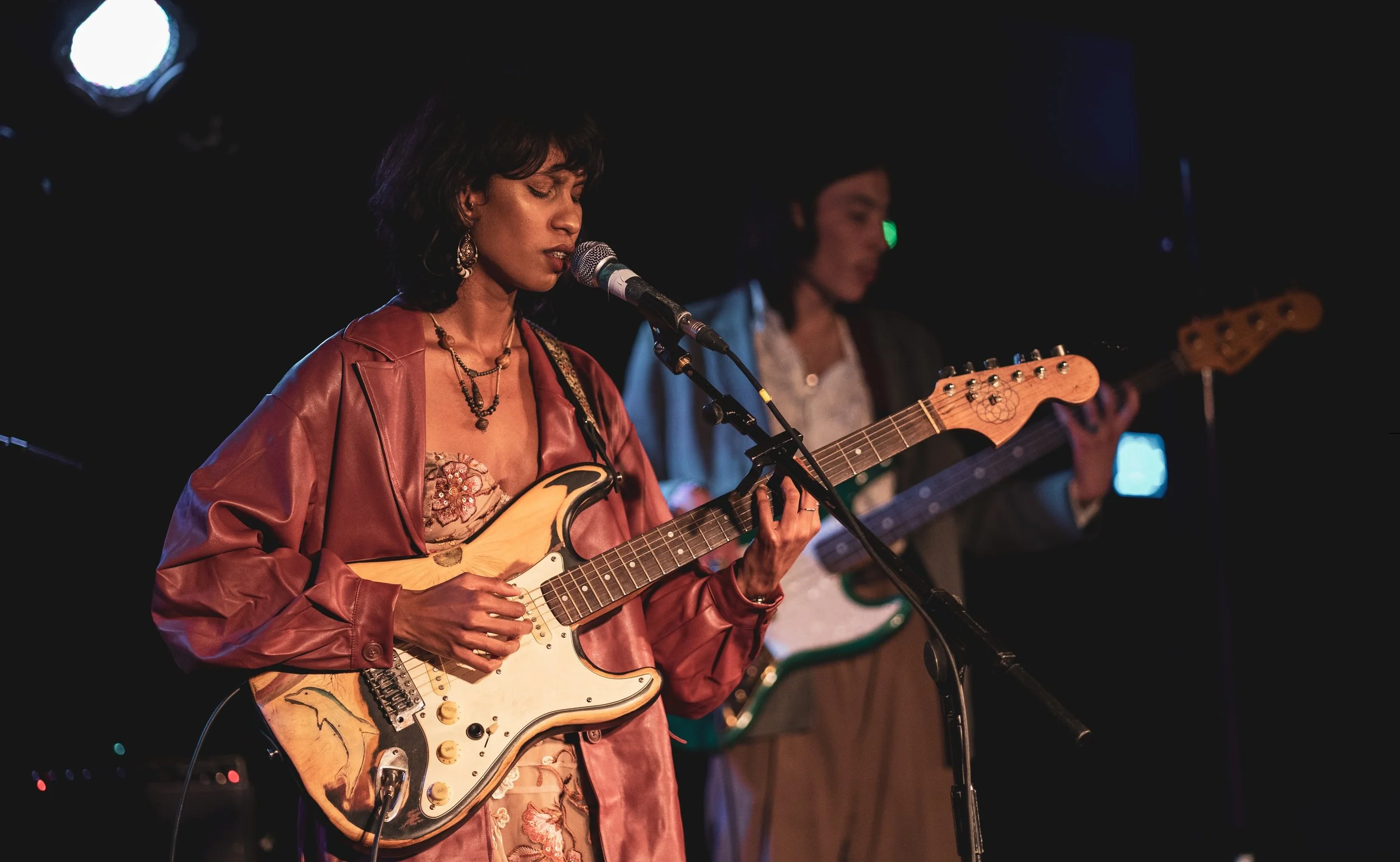A woman singing and playing an electric guitar on stage with another musician in the background.