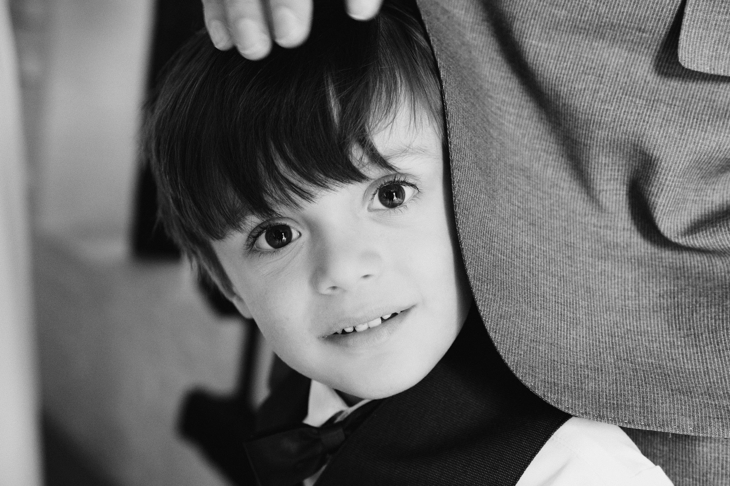 A young boy with dark hair and big eyes smiling, partially tilting his head and leaning against an adult. The adult wearing a grey jacket has one hand gently placed on the boy's head.