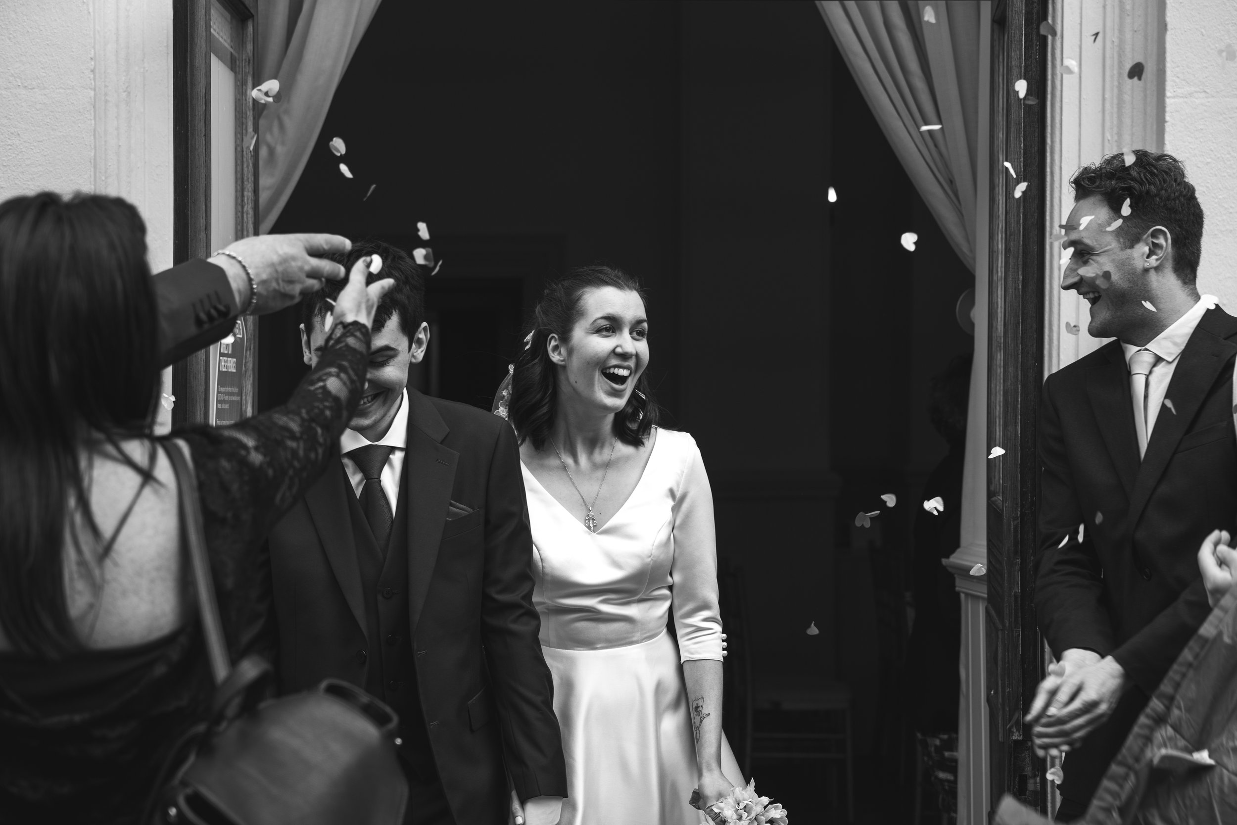 A black and white photo of a wedding scene with three laughing people. A woman in a wedding dress is at the center, smiling. Two men in suits are on either side, also smiling and engaging in conversation. Flower petals are falling around them.