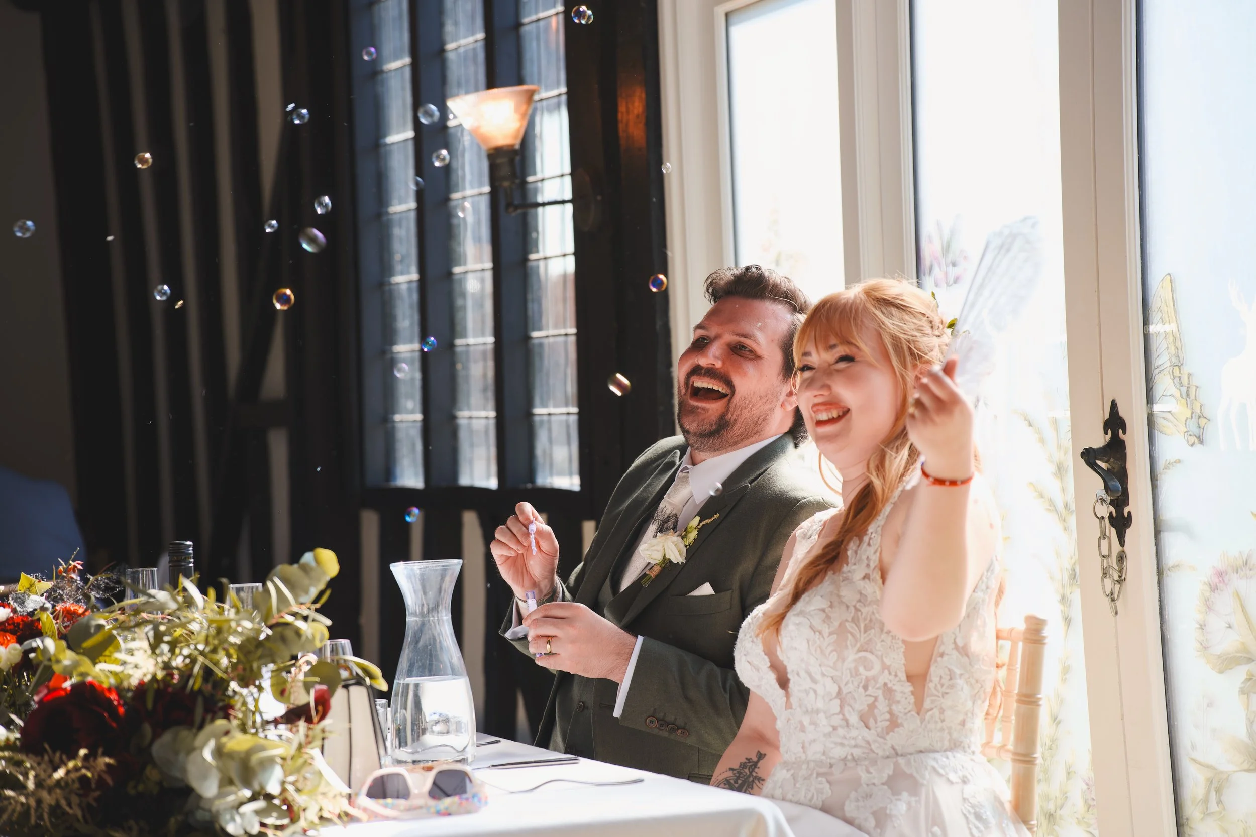 A man and a woman sitting at a table, smiling and enjoying a celebration with soap bubbles floating around, in a room with large windows and a black curtains backdrop.
