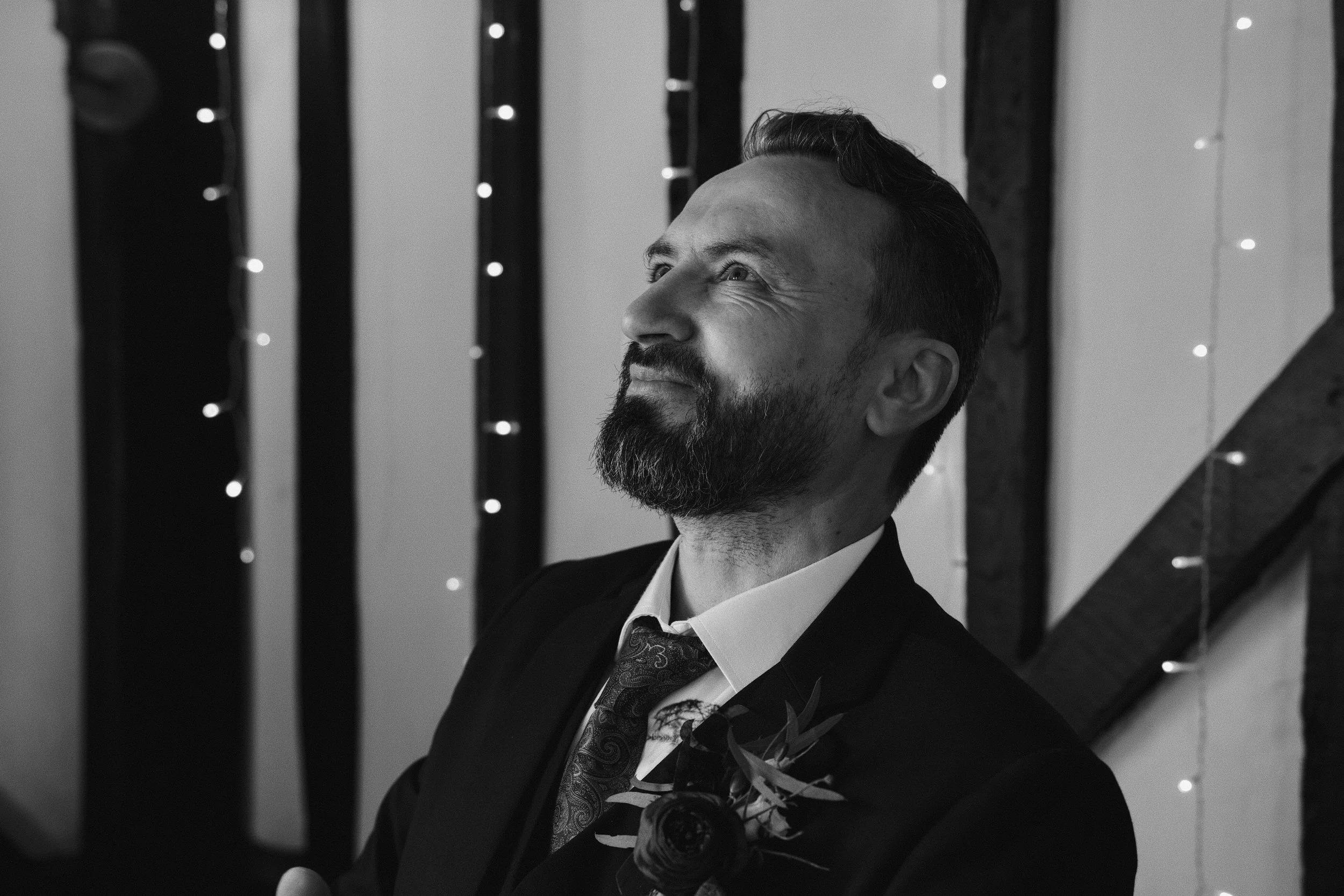 A black and white photo of a man with a beard and styled hair, dressed in a formal suit, smiling slightly, looking upwards. The background features vertical curtain-like stripes with small lights strung between them.