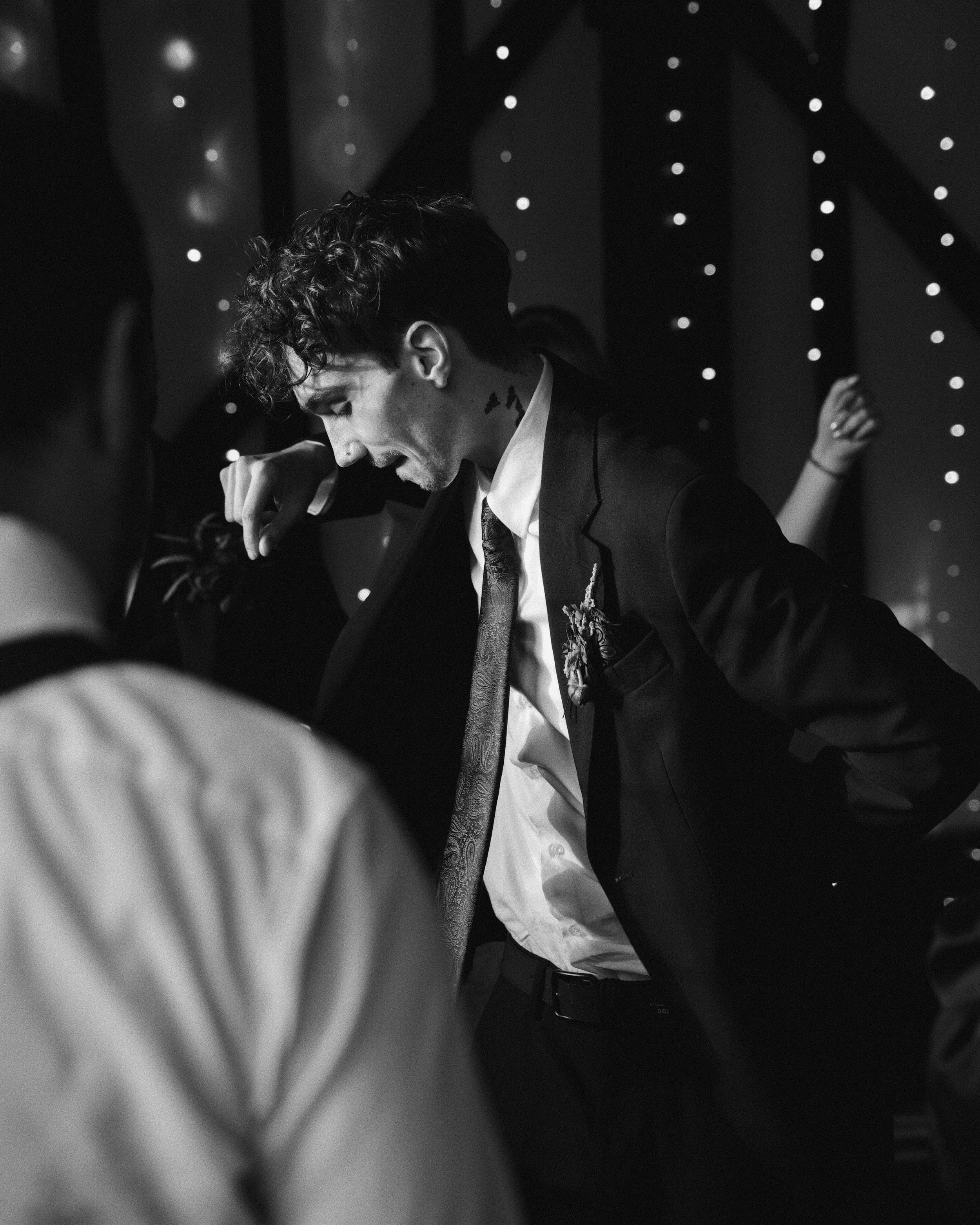 Black and white photo of a man in a suit with curly hair and tattoos on his neck, standing against a backdrop of small, bright lights. The image is slightly blurry and appears to be taken in a dimly lit environment.