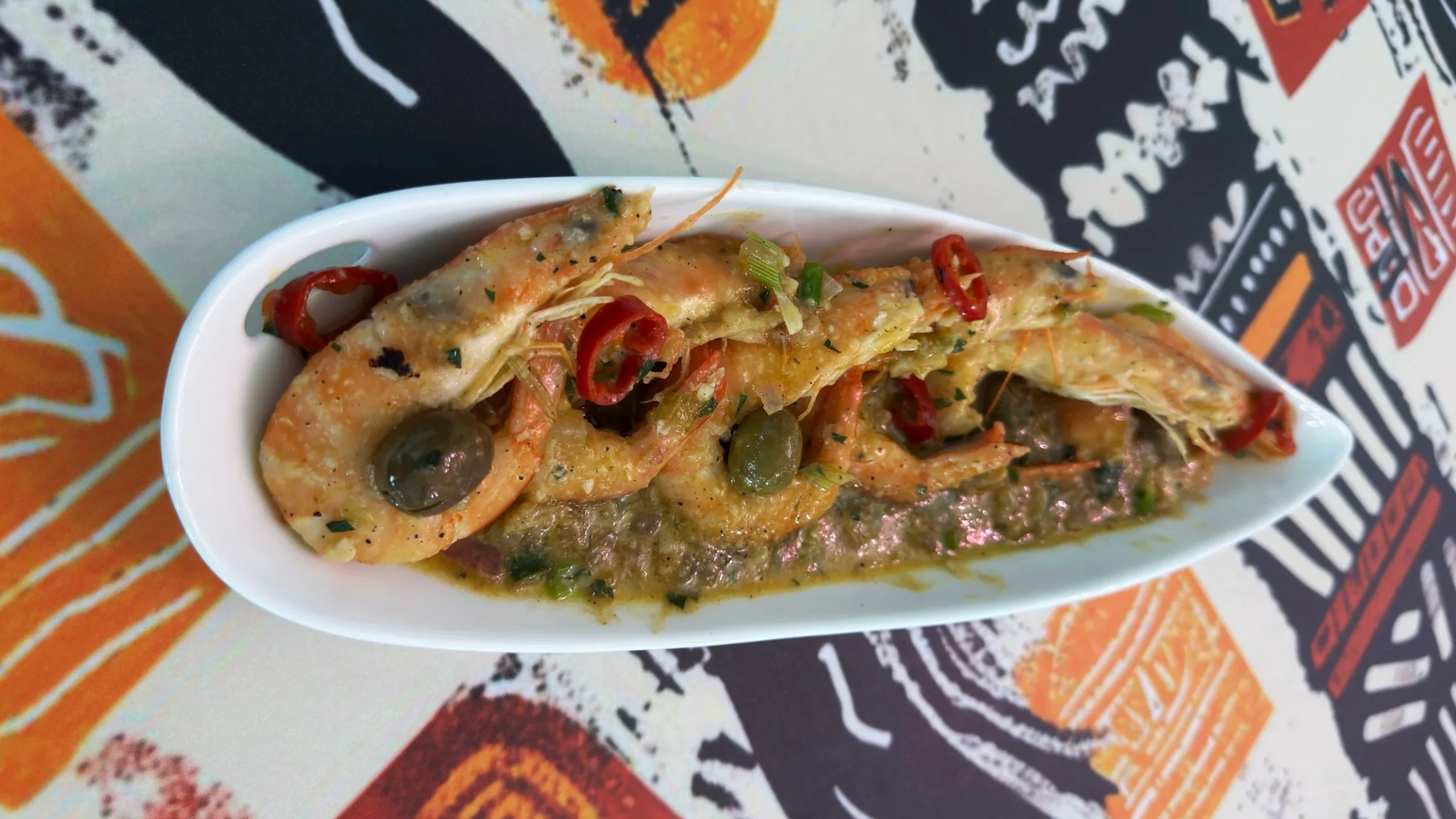 A small white bowl filled with a seafood dish containing shrimp, green and red chili peppers, olives, and chopped herbs, sitting on a colorful printed tablecloth.