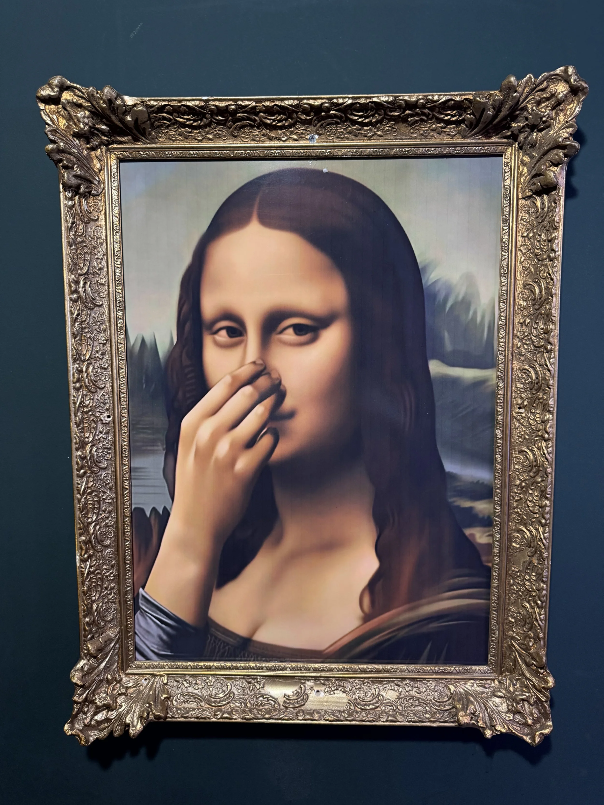A framed portrait of the Mona Lisa, holding fingers near her nose, with a background of a landscape featuring mountains and water.