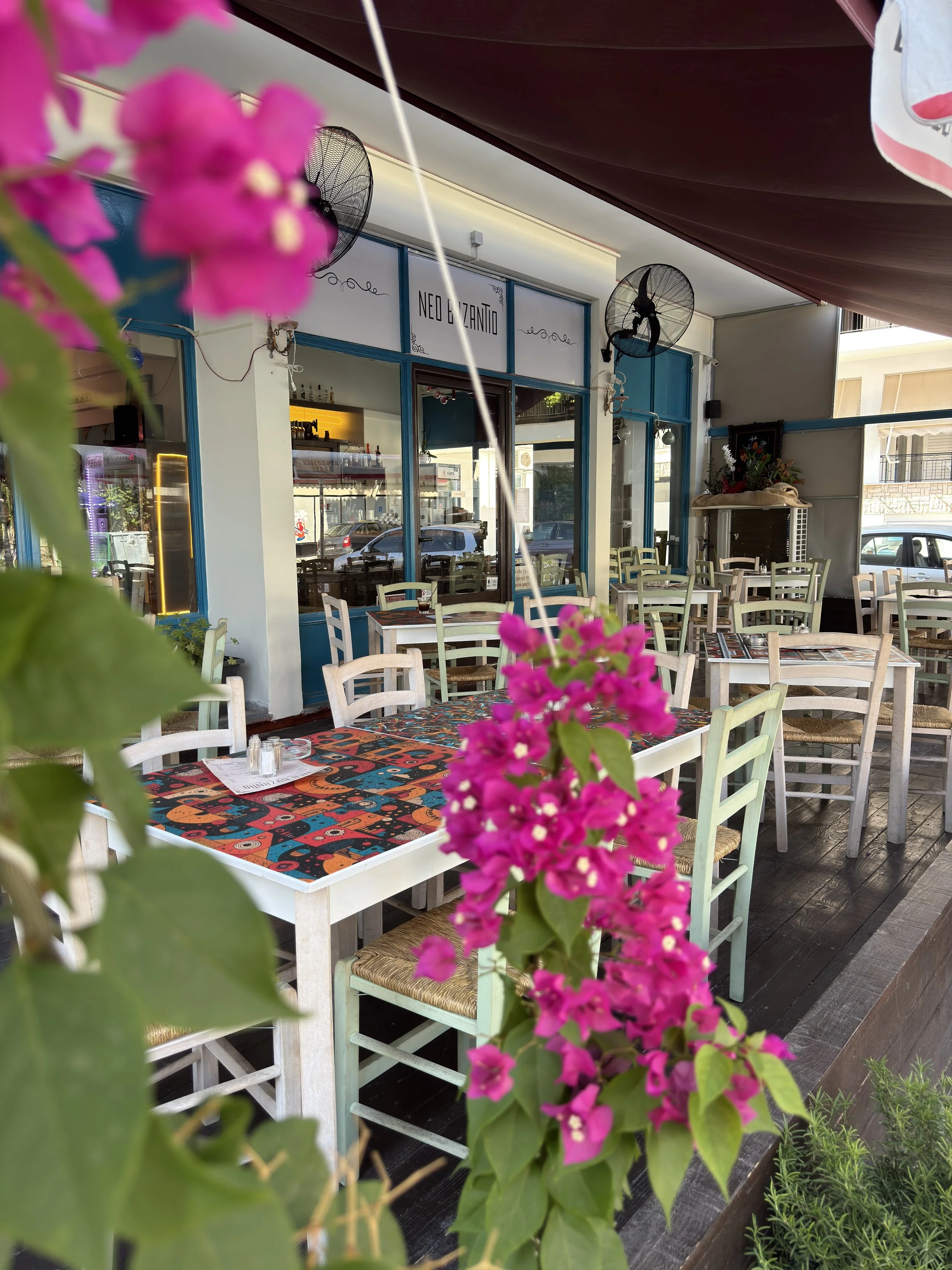 Outside seating area of a Neo Byzantio meze restaurant in Nafplio, with white chairs, colorful tablecloths, pink flowers in the foreground, and a sign reading 'NEO & BYZANTIO' on the wall.