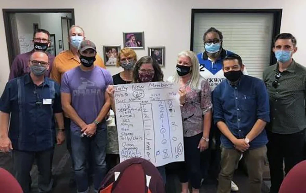 Group of ten people wearing masks, standing indoors, with a big sheet of paper that says "Tally New Member" and has voting tallies, in front of framed photos on the wall.