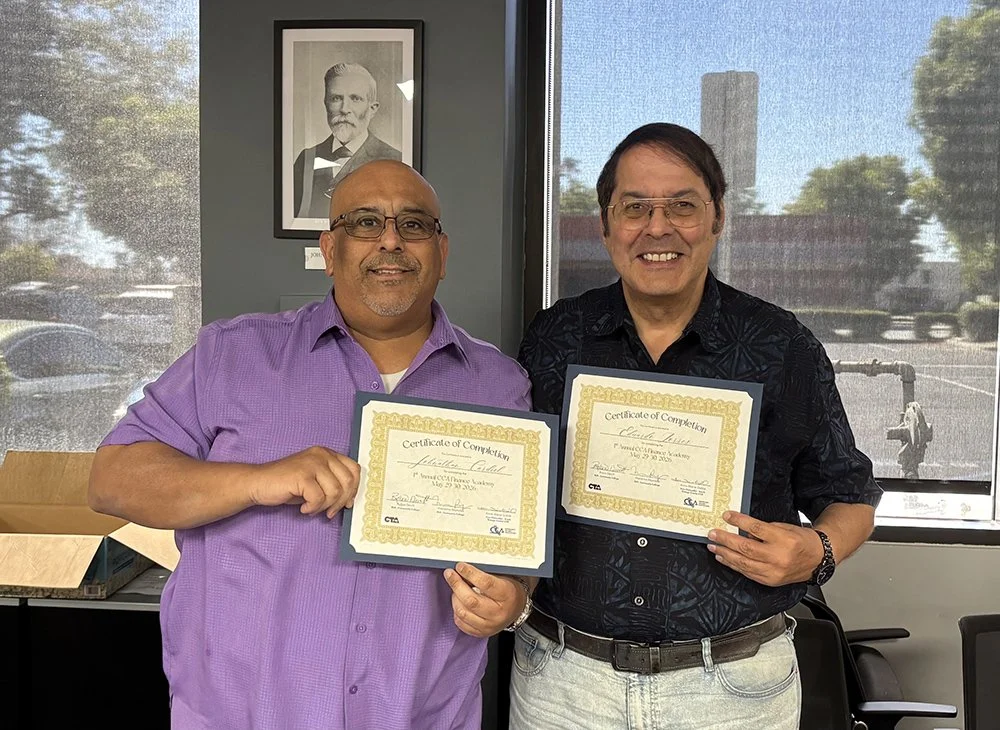 Two men standing inside an office, holding certificates of completion, smiling at the camera. Behind them is a window with a view of trees and parked cars, and a framed black-and-white portrait of a man with a beard on the wall.