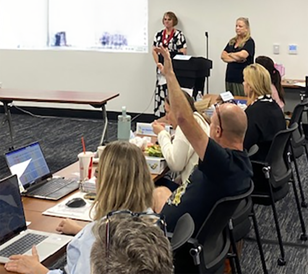 A group of people in a meeting room with laptops, some raising hands, and two women standing at the front near a whiteboard and a podium.