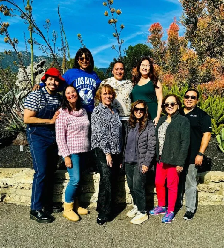 A group of nine people standing outdoors in front of a garden with various plants and trees, on a sunny day.
