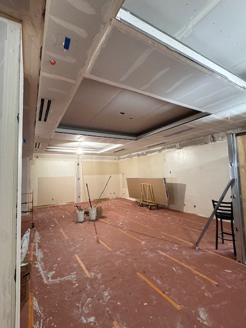 An unfinished room under construction with drywall, exposed ceiling framing, and construction materials on the floor.