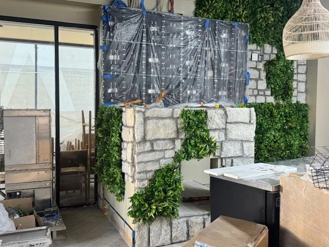 Indoor construction area with a stone fireplace surround, plastic sheeting for protection, plants, and various furniture and tools.