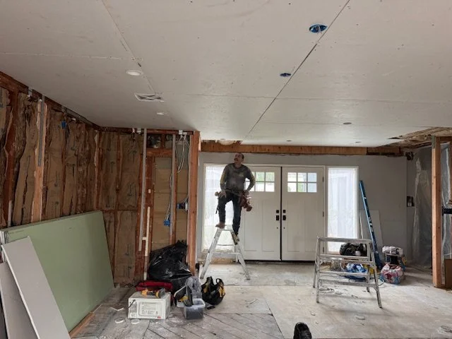 Construction worker standing on a ladder inside a partially renovated room with exposed walls. Building materials and tools are scattered around, including drywall panels leaning against the wall, a step ladder, and plastic bags. The room has a large