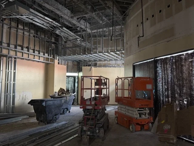 Construction site with scaffolding, construction lifts, waste bins, and exposed ceiling pipes and wires inside a building under construction.