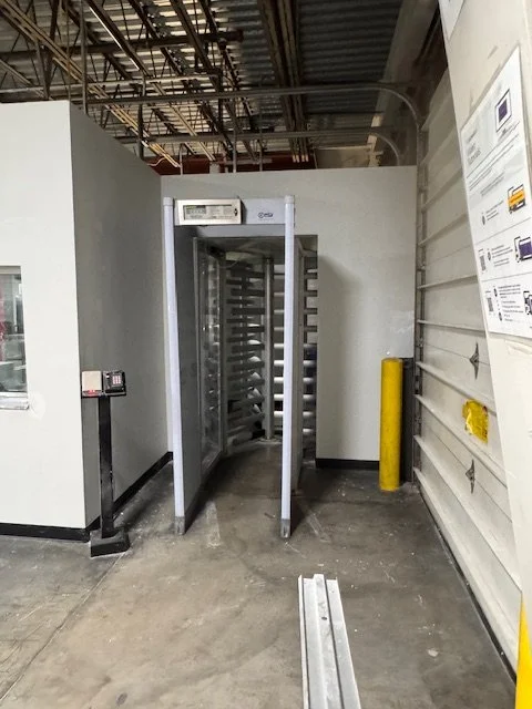 A walk-through metal detector in a warehouse or loading dock area, with industrial ceiling and walls, and a yellow safety post nearby.