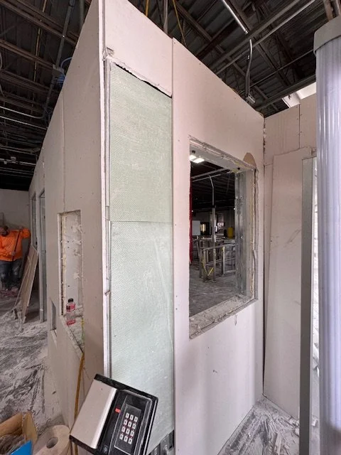 Interior of a construction site showing an unfinished room with drywall, a large window opening, and construction tools, including a phone on a stand, with exposed ceiling and piping.