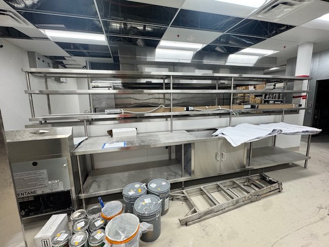 Empty stainless steel kitchen prep station with shelves, buckets of paint, a ladder, and some white cloths, in a commercial kitchen or food prep area.