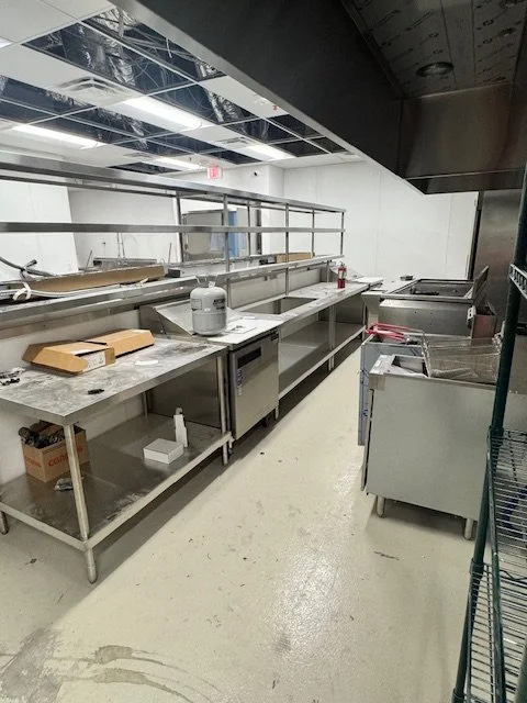 Empty industrial kitchen prep area with stainless steel counters and equipment, some pizza boxes on the counters, and an unfinished ceiling with exposed framing.