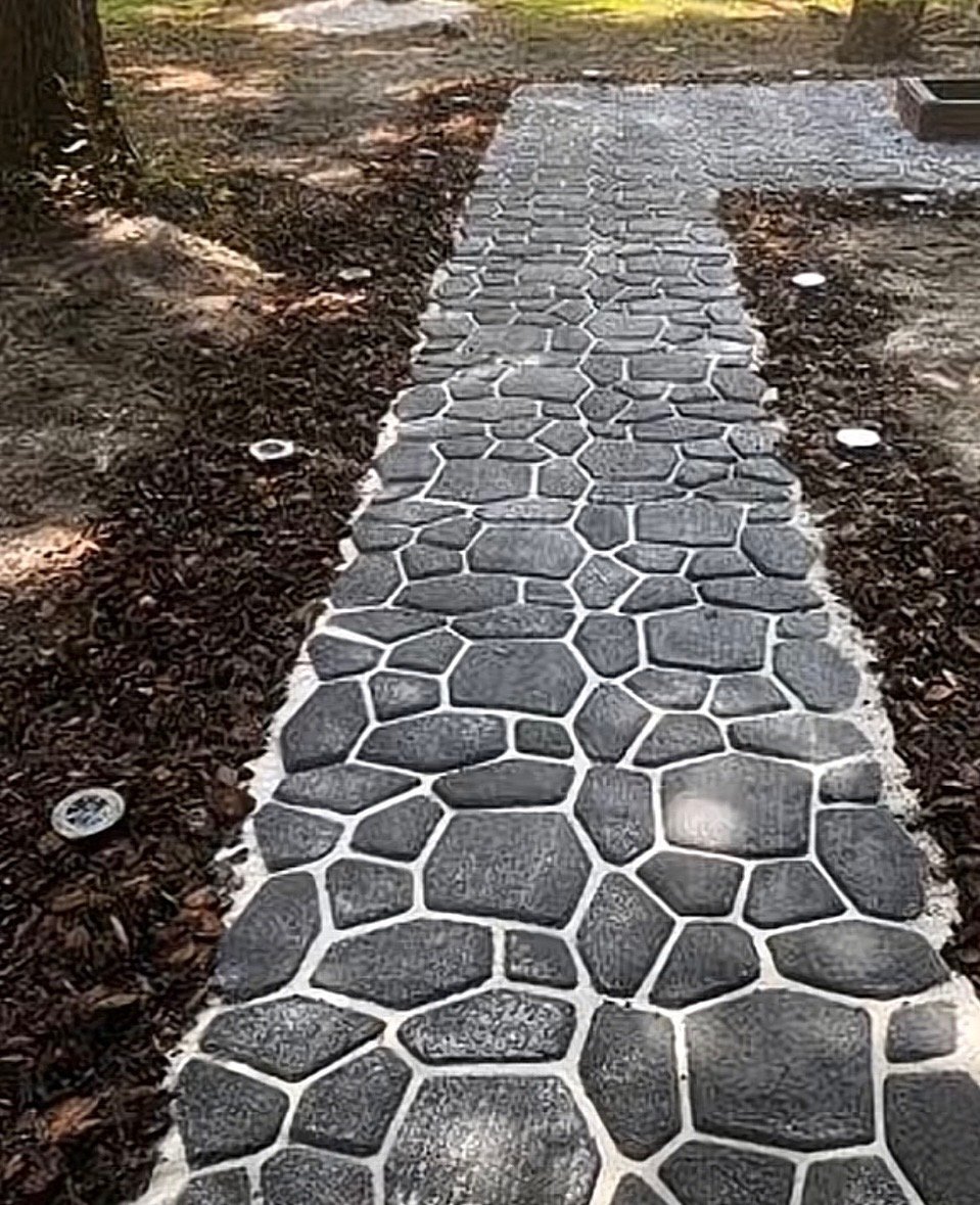 A stone pathway made of irregularly shaped dark stones with white grout, set in a yard with dirt and mulch on sides, and some outdoor lights or fixtures along the edges.