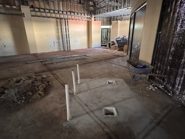 Construction site with exposed walls, concrete floor, and plumbing pipes. Tools and materials are scattered around.
